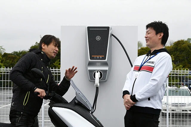 青木氏(左)の直撃に「これまでの50cc相当のEVから一歩先を行く本格派を造りたかった。静かで振動が少ないEVは疲れず移動できるので通勤の足に最適」と語る開発責任者の丸尾氏(右)