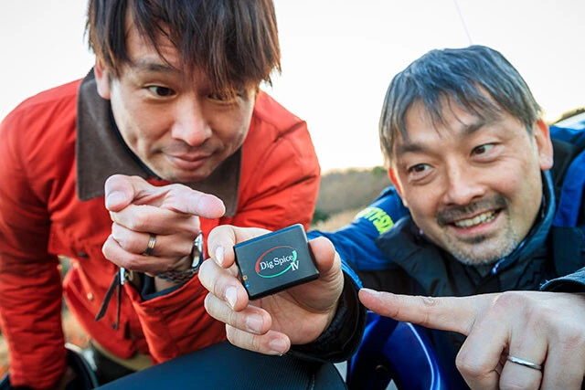 『ヤングマシン』の松田編集長（右）から時速60キロ到達タイムを計る乗り役に指名されたアオキ（左）は、取材の秘伝アイテム、モータースポーツ用データロガー（記録機器）「デジスパイスⅣ」を託された