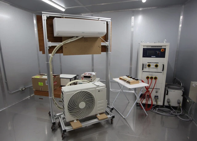 ウオークインチャンバー（恒温恒湿室）でのエアコン性能テスト。さまざまな温度・湿度環境での耐久性とエネルギー消費特性を確認する。省エネ性能の検証に不可欠