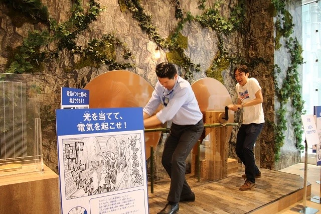 金狼と銀狼が千空が作った「人力発電機」を使うシーンを再現し、中央のランプを点灯させるＡＧＣ社員のふたり。特に奥の小長谷さんは金狼のように必死な表情を作るほどノリノリ　©米スタジオ・Ｂｏｉｃｈｉ／集英社