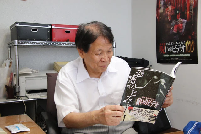 2017年に刊行した『ほんとにあった! 呪いのビデオ 恐怖のヒストリー』を懐かしそうに眺める張江さん。同書には74作目までのデータベースや歴代監督のインタビューが掲載されている。ちなみに張江さん自身はこれまで「本当の裏方なので」とほとんど表舞台に出てこなかった