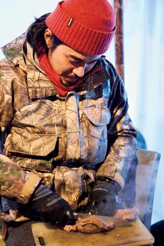 ヒザの上にまな板を置いて、愛犬用のごはんのシカ肉を切る東出。山小屋というインドアにいるけど、アウトドア料理!?