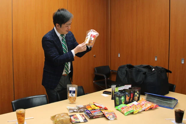 パッケージを愛おしそうに見つめながら、説明する中倉氏。ちなみに奥のバッグいっぱいに柿ピーが入っていた