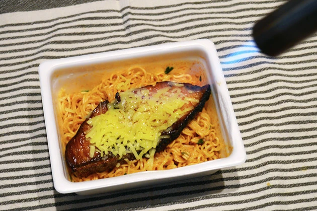 (3)バーナー! (2)を耐熱皿に移し、ブリの照り焼きとチーズをのせてガスバーナーであぶる。レンチンでもOK。今回は容器を移さずあぶったらカップが溶けてしまった。絶対にマネしないこと!