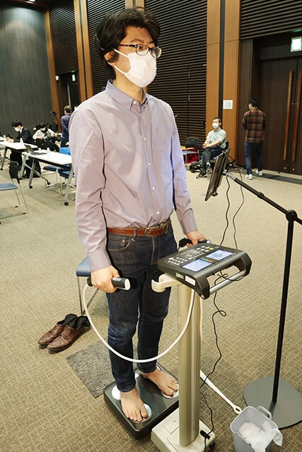 はだしで体組成計に乗って両手でグリップを握る編集・カワサキ