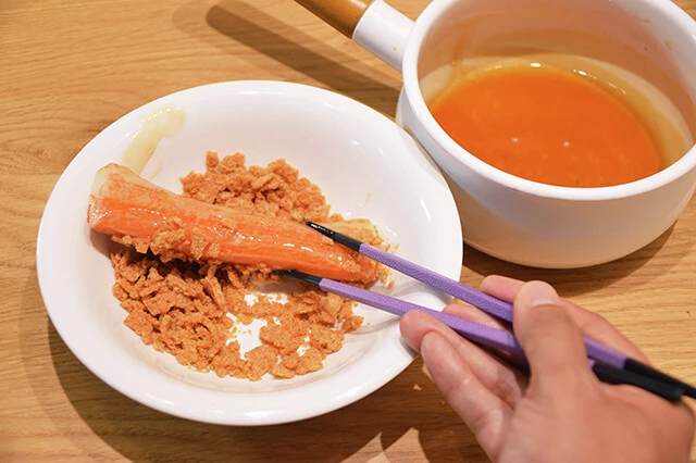(3)つける! カニカマスティックを(1)の食器に入れ、粉々になったスナックをまぶして全体にたっぷりと付着させる。海老天の衣のようになったら盛りつけ皿に移そう