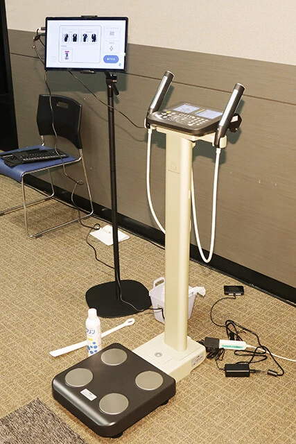 左に設置されたタブレットとタニタの体組成計を組み合わせた機器を利用する今回のサービス。体組成計は新開発されたものではなく業務用として販売されている従来のモデル