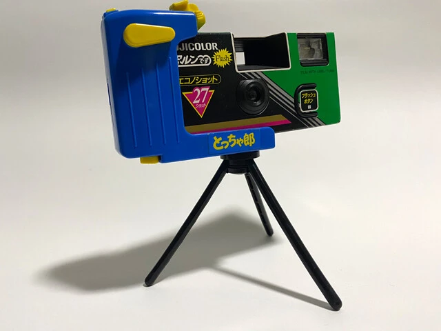 「とっちゃ郎」はレンズ付きフィルムにセルフタイマーを外付けする三脚。左側だけを挟み込む機構と、可変のシャッターボタンによっていろいろな機種に対応するようになっている。この「写ルンです エコノショットFlash27」は平成4年発売