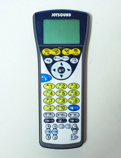 通信カラオケのリモコンと目次本。目次本には楽曲名と対応する番号が並んでいる。番号をリモコンで本体に送信して曲データを呼び出す