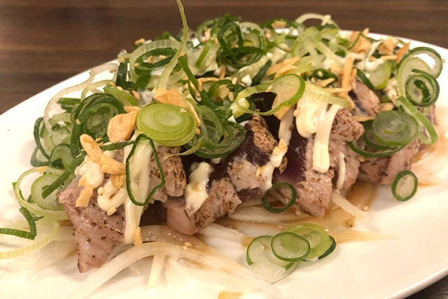 「鉄板居酒屋 丸一」の「カツオのたたきwithマヨネーズ」。マヨの程よい酸味と油がカツオの臭みを消し、トロのようなうまみを感じさせる。ただしネギが多すぎるのか、全部口に入れると少々辛い