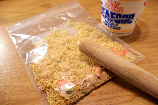 （１）砕く！　シーフードヌードルの中身をすべて取り出してジップロックなどに移し、すりこぎなどを使って全体を叩き、麺を細かく粉砕する。ベビースターラーメンを砕くイメージで叩こう