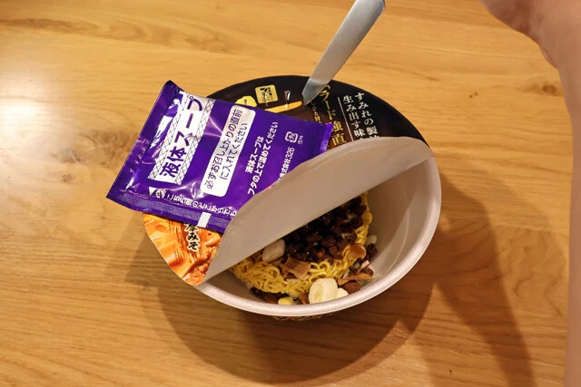 （２）開ける！　セブンプレミアムのカップ麺「すみれ 濃厚札幌みそ」のフタに包丁などで湯切り穴を作り、カップの中からスープ2袋を取り出す。かやくだけ入れて熱湯を注ぎ、5分待とう