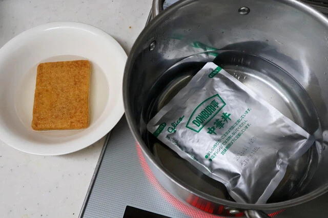 （１）湯煎！　お湯を沸騰させ、レトルト中華丼の具を温める。同時にどん兵衛のお揚げを食器に移し、熱湯を注いで戻す。お揚げは柔らかく戻ったら水気を軽く切り、ひと口大にカットする