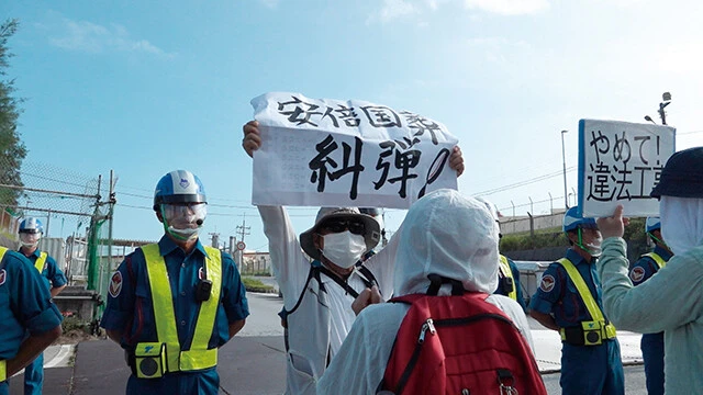 辺野古の米軍新基地建設現場にて、抗議活動を行なう人々。「安倍国葬糾弾!」「やめて!違法工事」といった文字が掲げられている。©「国葬の日」製作委員会