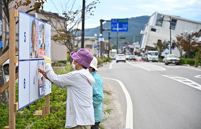 輪島市でポスターを張る選挙陣営の関係者たち。右奥には地震の影響で横倒しになった五島屋ビルが見える