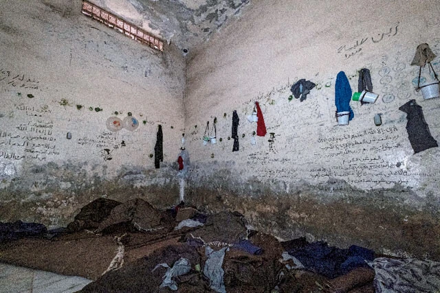 アサド政権によってテロ容疑をかけられた市民が収容されていたという、メッゼ空軍基地内の収容所。収容者は光のほとんど届かない劣悪な環境に閉じ込められていた。石鹸で作ったフックにかけられた衣類や日用品から、その過酷な生活の一端が想像できる。壁には勾留された人々が神に救いを求めるメッセージを残していた